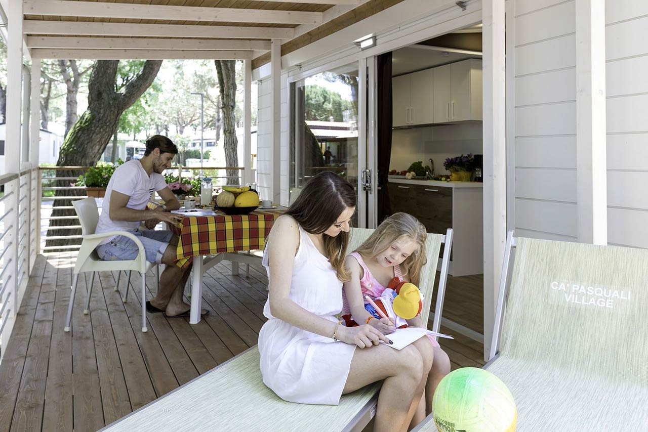 In primo piano mamma e bambina colorano su un lettino nella veranda di una Mobile Home. Sullo sfondo il papà legge.