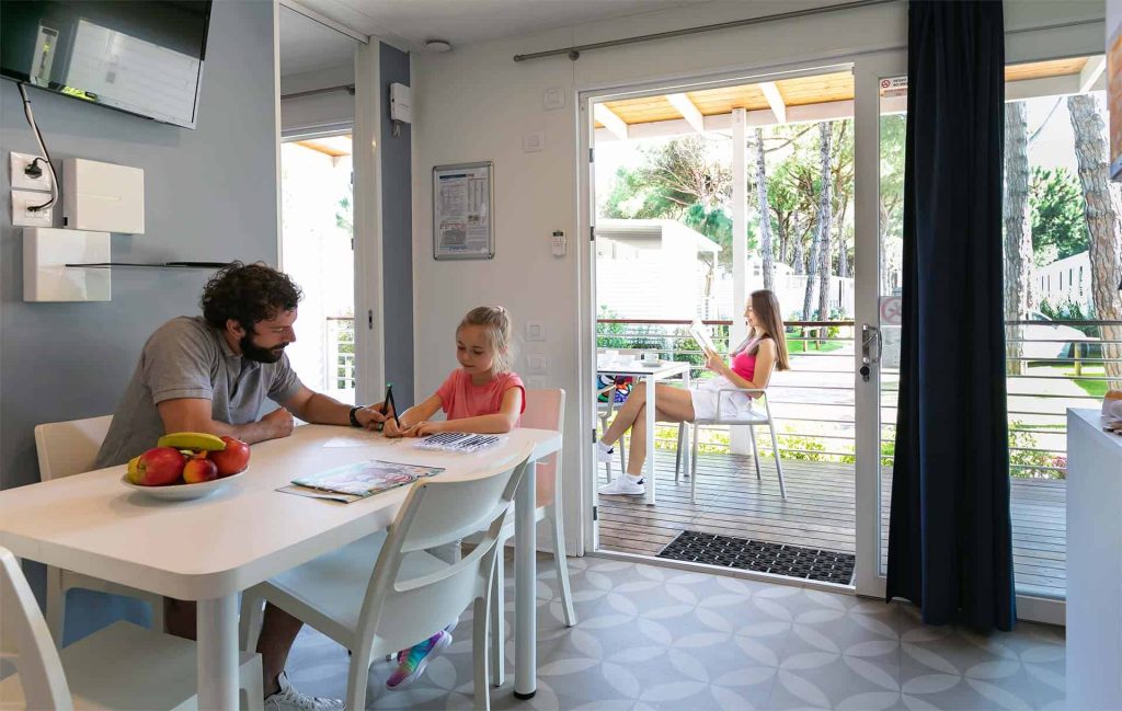 In primo piano papà e figlia che colorano nel soggiorno di una Mobile Home. Sullo sfondo la mamma legge una rivista nella veranda.